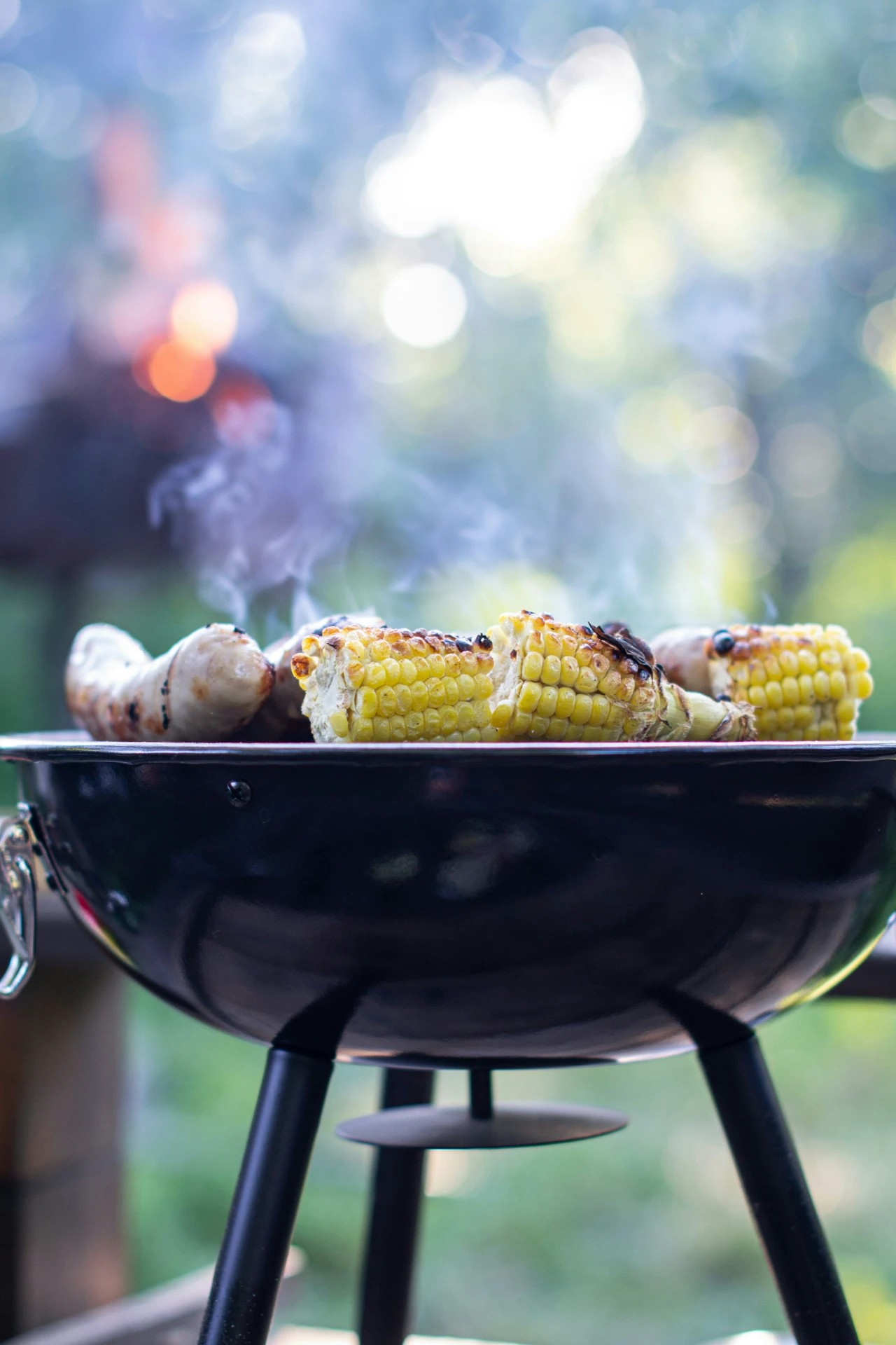 Close-up of corn and sausages grilling on a portable barbecue grill with a blurred outdoor fire in the background, perfect for a private chef BBQ party experience.