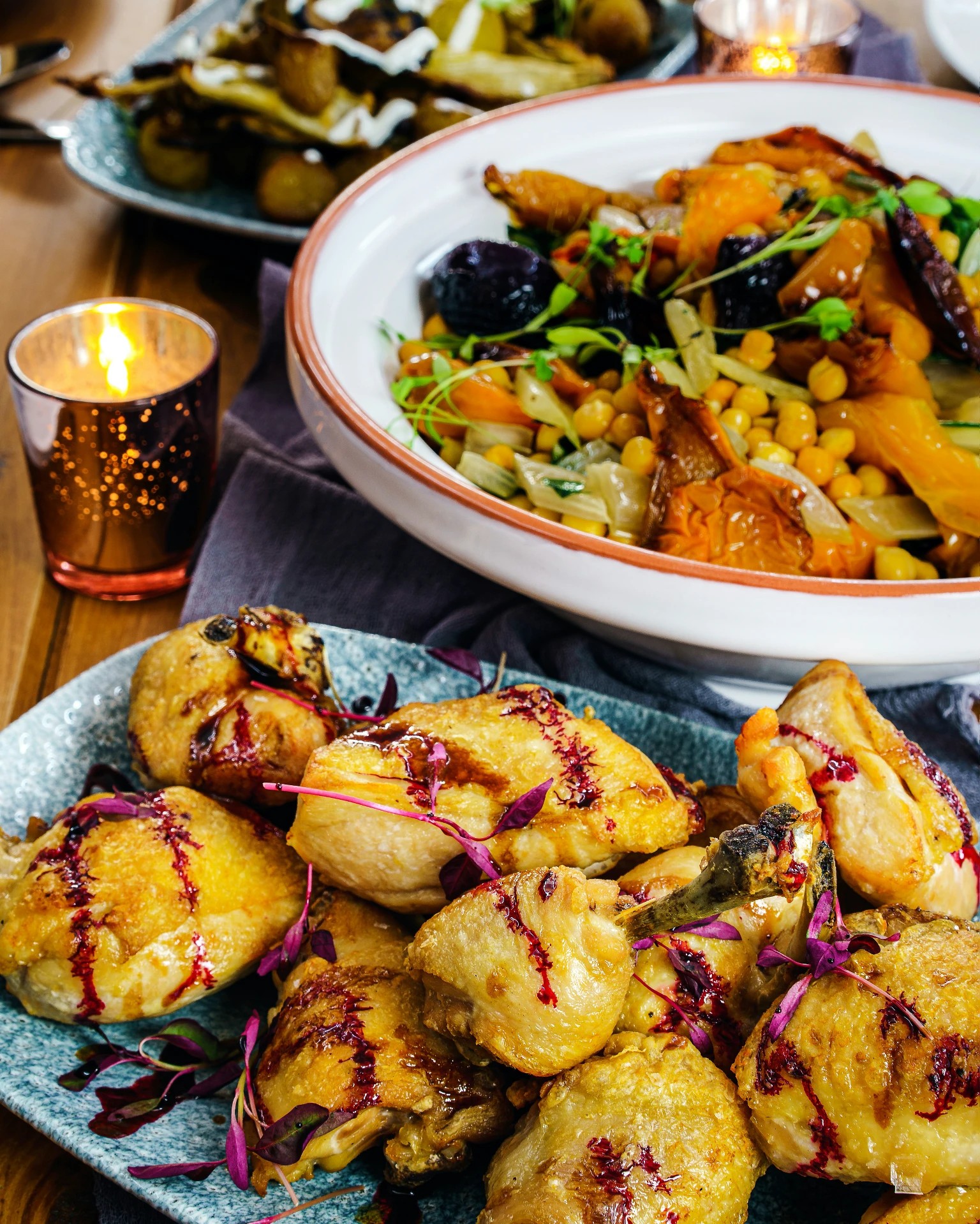 Close-up of roasted chicken drizzled with berry reduction, paired with a bowl of roasted vegetables and chickpeas, set on a candlelit dining table for an intimate private chef BBQ party.